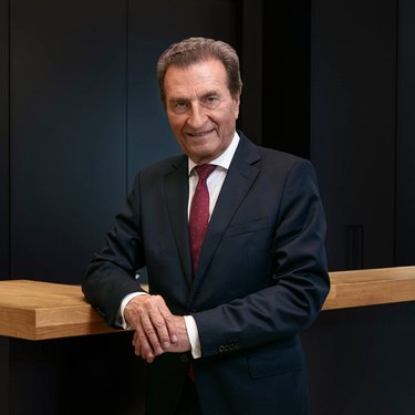 A man in a suit stands at a wooden bar, smiling, with a modern backdrop in dark colors.