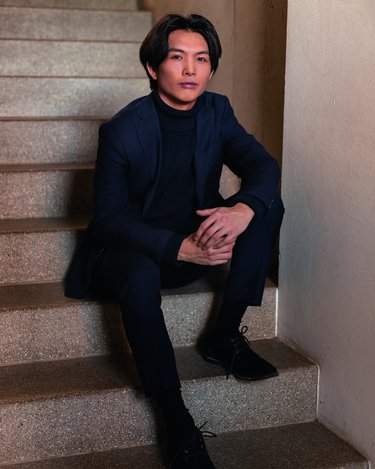 Ein Mann sitzt auf einer Treppe, trägt einen dunklen Anzug und einen Rollkragenpullover. Er hat mittellange Haare und schaut direkt in die Kamera.