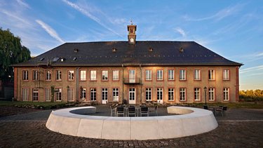 Ein historisches Gebäude mit einem grauen Dach und einer Uhr im Giebel, umgeben von einem runden, weißen Sitzbereich. Die Fassade hat große Fenster und eine symmetrische Anordnung. Der Himmel ist klar mit einigen Wolken.