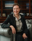 Eine Frau in einem grauen Anzug sitzt entspannt auf einem Ledersofa und lächelt in die Kamera. Bücherregale sind im Hintergrund sichtbar.