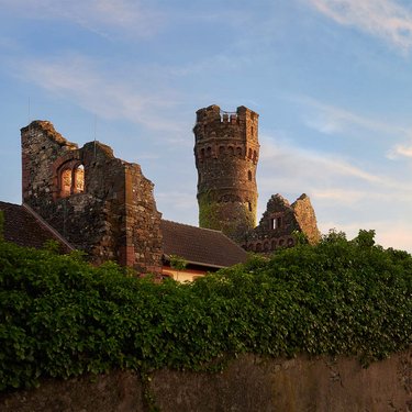 Eine teilweise verfallene Burg mit einem hohen Turm, umgeben von üppigem Grün und einem klaren Himmel im Hintergrund. Die Struktur zeigt historische Architektur und ist von einer Mauer umgeben.