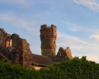 Eine teilweise verfallene Burg mit einem hohen Turm, umgeben von üppigem Grün und einem klaren Himmel im Hintergrund. Die Struktur zeigt historische Architektur und ist von einer Mauer umgeben.
