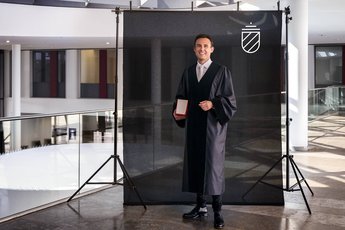 Ein Mann in einem schwarzen Talar steht vor einem schwarzen Hintergrund und hält ein Buch in der Hand. Er trägt ein weißes Hemd und eine Krawatte und lächelt in die Kamera.