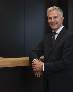 A man in a suit stands at a wooden bar and smiles. The background is dark and modern in design.