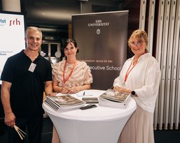 Drei Personen stehen an einem Tisch mit Informationsmaterialien über die EBS Universität und die Executive School.