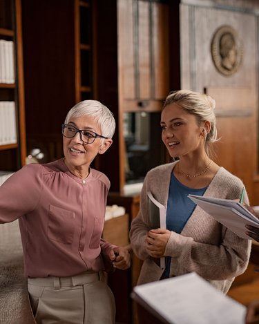 Eine ältere Frau mit kurzen, grauen Haaren erklärt etwas an einem Flipchart, während drei jüngere Personen aufmerksam zuhören. Die Umgebung ist eine Bibliothek mit Regalen voller Bücher.