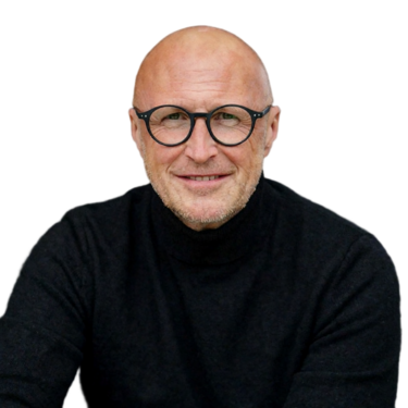 Mann mit Glatze und Brille, trägt einen dunklen Anzug und ein weißes Hemd, steht vor einem neutralen Hintergrund.