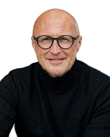 Mann mit Glatze und Brille, trägt einen dunklen Anzug und ein weißes Hemd, steht vor einem neutralen Hintergrund.
