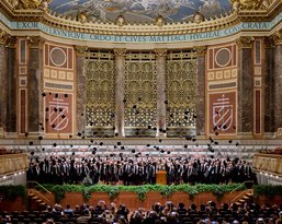 Ein großer Chor steht auf der Bühne eines prächtigen Konzertsaals, umgeben von aufwendiger Architektur und Dekoration.