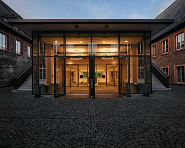 Moderne Glastüren führen in einen hell erleuchteten Innenraum, umgeben von historischen Gebäuden mit roten Fensterrahmen. Der Boden besteht aus gepflasterten Steinen, die den Eingangsbereich umgeben.