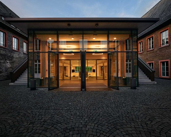 Moderne Glastüren führen in einen hell erleuchteten Innenraum, umgeben von historischen Gebäuden mit roten Fensterrahmen. Der Boden besteht aus gepflasterten Steinen, die den Eingangsbereich umgeben.