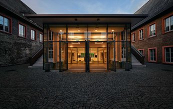 Moderne Glastüren führen in einen hell erleuchteten Innenraum, umgeben von historischen Gebäuden mit roten Fensterrahmen. Der Boden besteht aus gepflasterten Steinen, die den Eingangsbereich umgeben.