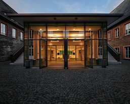 Moderne Glastüren führen in einen hell erleuchteten Innenraum, umgeben von historischen Gebäuden mit roten Fensterrahmen. Der Boden besteht aus gepflasterten Steinen, die den Eingangsbereich umgeben.