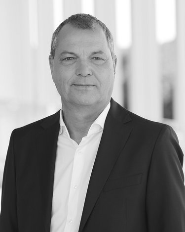 Ein Mann in einem schwarzen Anzug mit weißem Hemd steht vor einer modernen Architektur mit Säulen im Hintergrund.