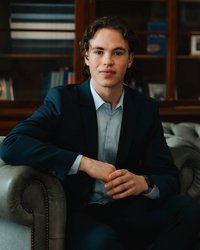 A young man in a dark blue suit sits relaxed on a gray sofa, looking directly at the camera.