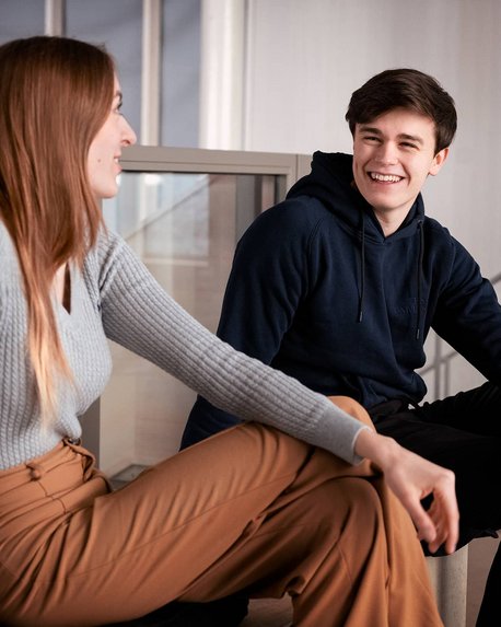 Two young adults are sitting in a modern room, smiling as they chat. The woman is wearing a gray sweater and beige pants, while the man is in a blue hoodie.