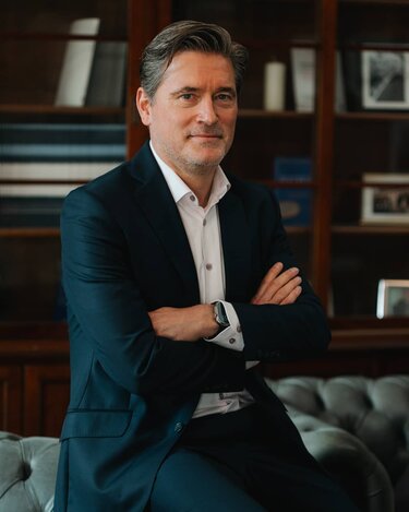 Ein Mann in einem dunklen Anzug sitzt mit verschränkten Armen auf einem Sofa. Im Hintergrund sind Bücherregale sichtbar.