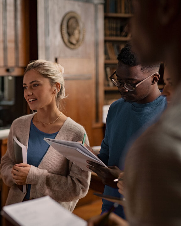 An older woman is explaining to a younger woman and a man in front of a whiteboard, with bookshelves in the background.