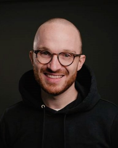 A man with a bald head and glasses smiles at the camera. He is wearing a black hoodie and has a short, well-groomed beard. The background is dark and neutral.
