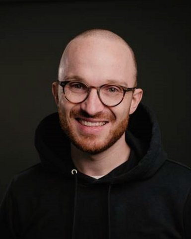Ein Mann mit Glatze und Brille lächelt in die Kamera. Er trägt einen schwarzen Kapuzenpullover und hat einen kurzen, gepflegten Bart. Der Hintergrund ist dunkel und neutral.