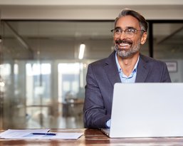 Ein lächelnder Mann mit Brille sitzt an einem Tisch vor einem Laptop, umgeben von Büromaterialien in einem modernen Büro.