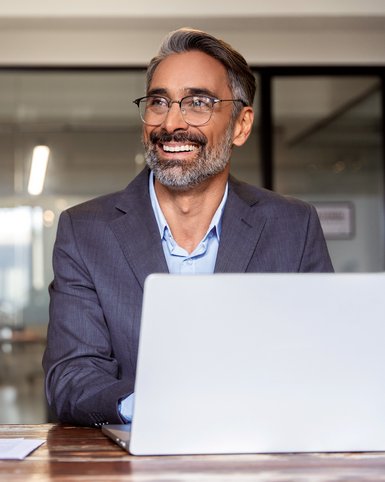 Ein lächelnder Mann mit Brille sitzt an einem Tisch vor einem Laptop, umgeben von Büromaterialien in einem modernen Büro.